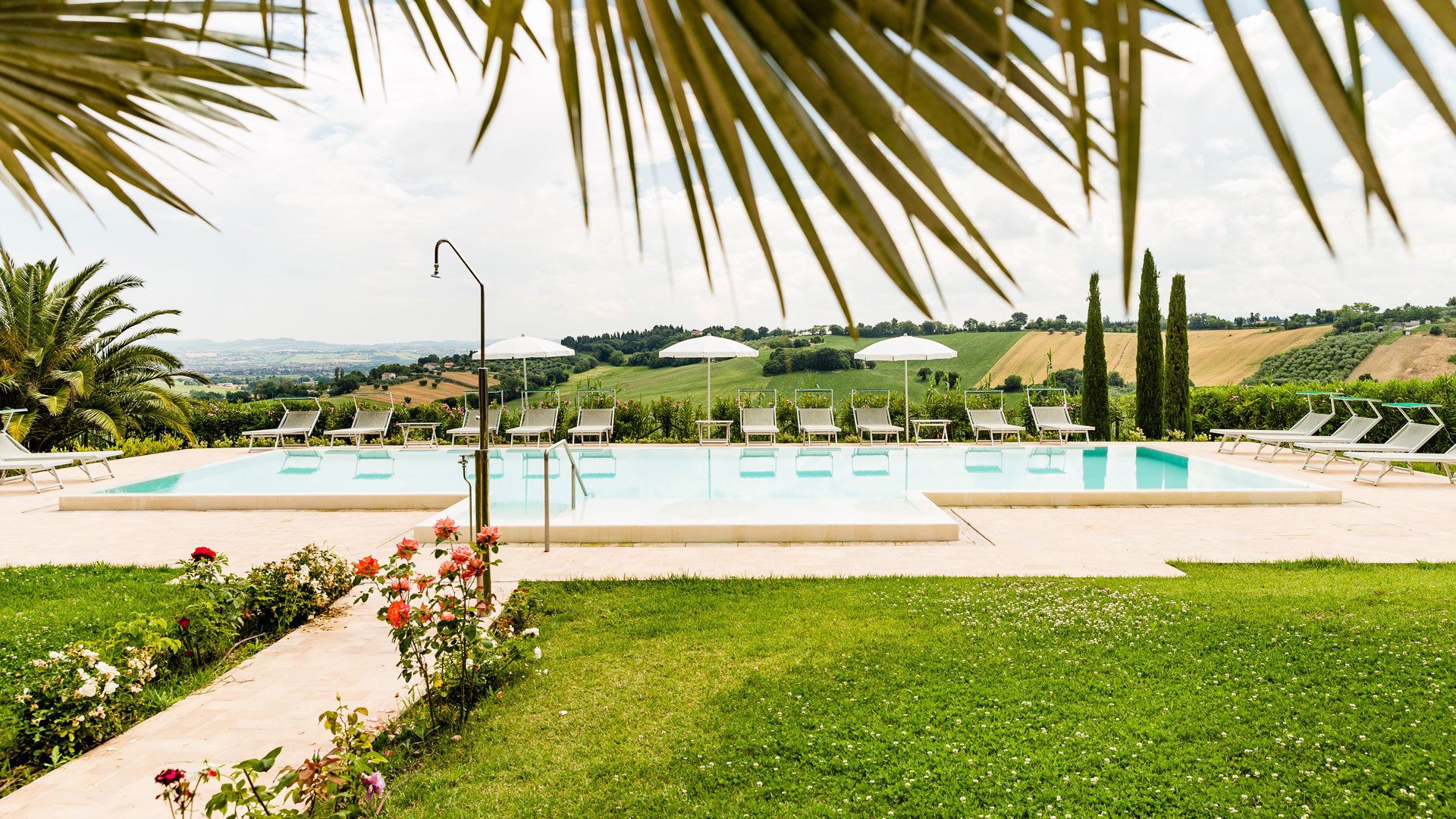 A pool area with multiple lounge chairs and umbrellas, surrounded by greenery and a view of rolling hills.