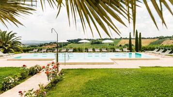 A pool area with multiple lounge chairs and umbrellas, surrounded by greenery and a view of rolling hills.