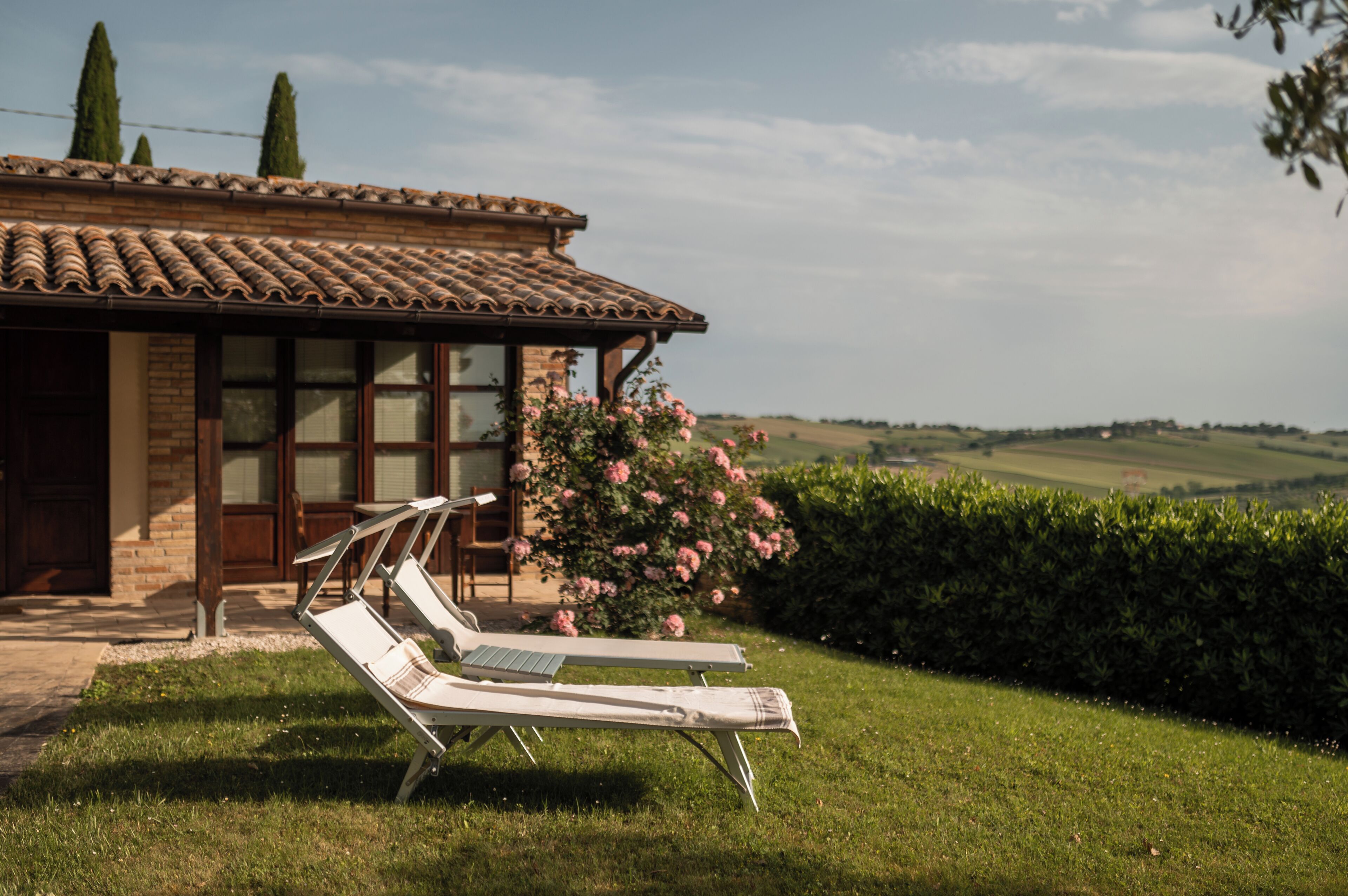 A villa with a tiled roof, a brick facade, and a garden with two lounge chairs and blooming pink flowers.