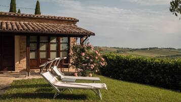 A villa with a tiled roof, a brick facade, and a garden with two lounge chairs and blooming pink flowers.