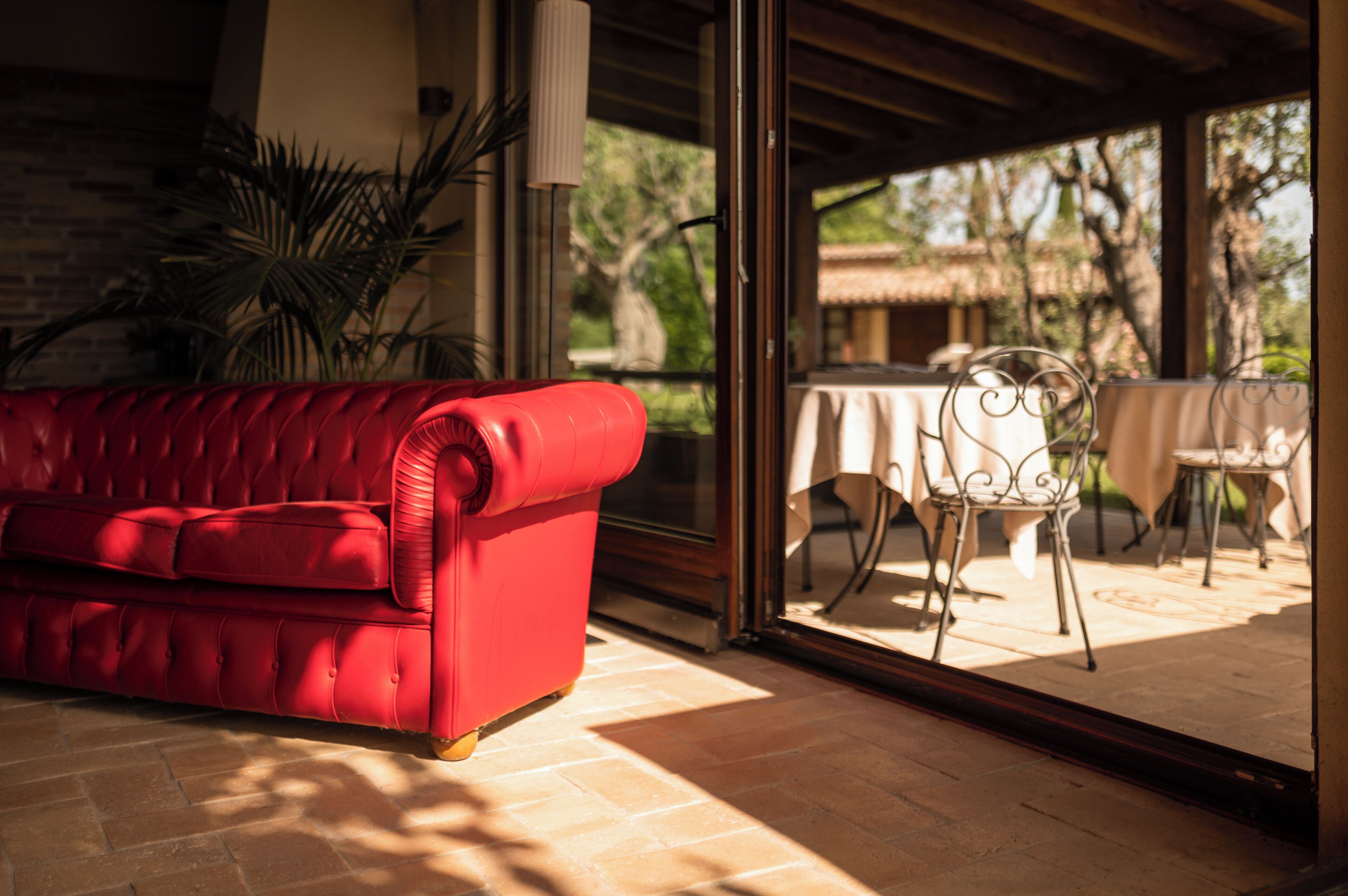 A red sofa in a sunlit room with a glass door leading to an outdoor patio area with a table and chairs.