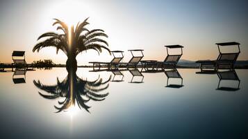 A serene beach scene with a palm tree and sunlit lounge chairs reflecting on the water.