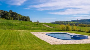 Una piscina con borde de piedra, rodeada por un césped y árboles, con un paisaje montañoso de fondo.