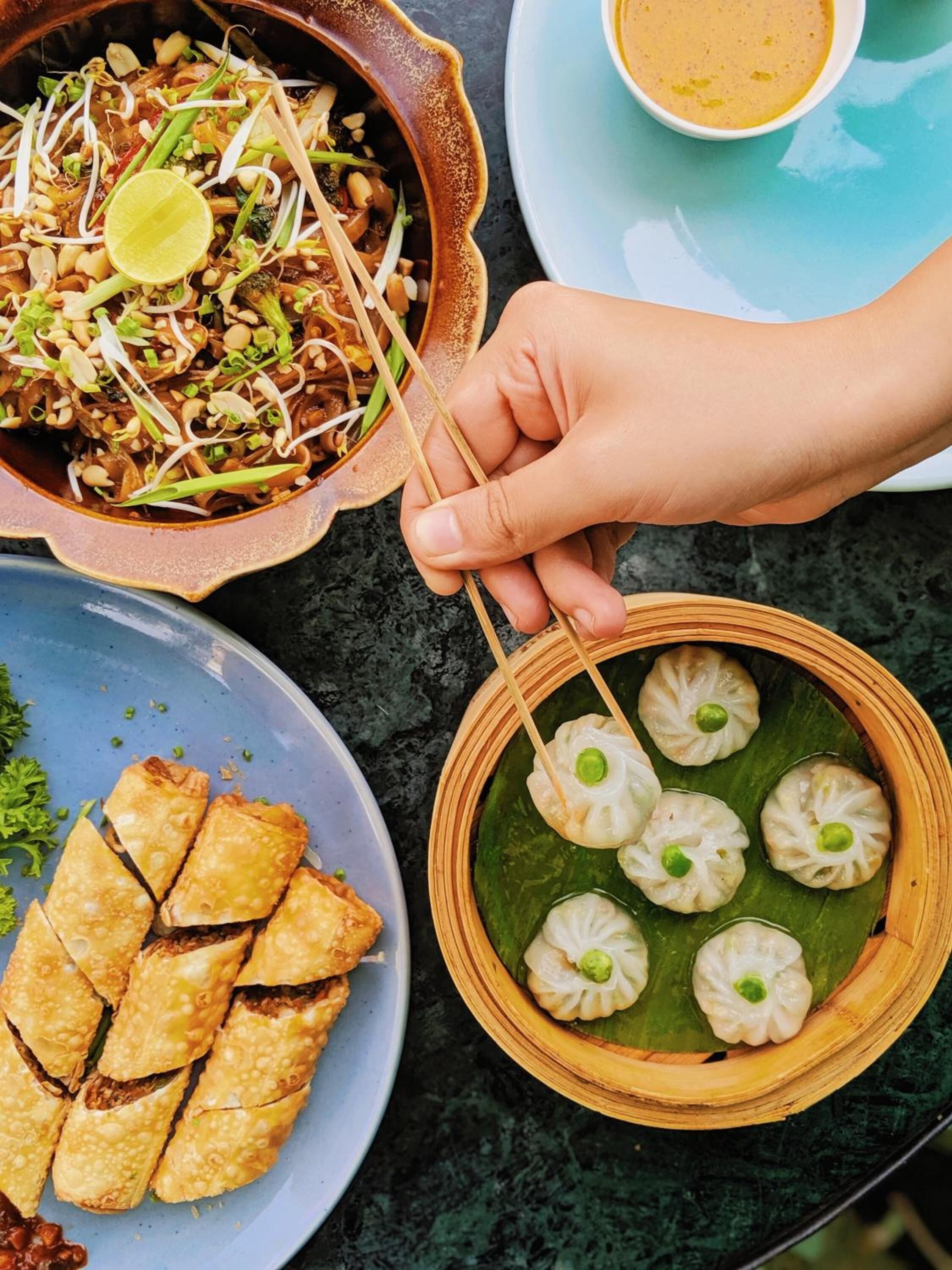 A meal with various dishes including a bowl of noodles, a plate of spring rolls, and a basket of dumplings.