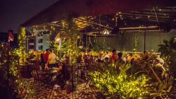 Un área de comedor al aire libre con mesas y sillas, rodeada de plantas en macetas y luces colgantes.