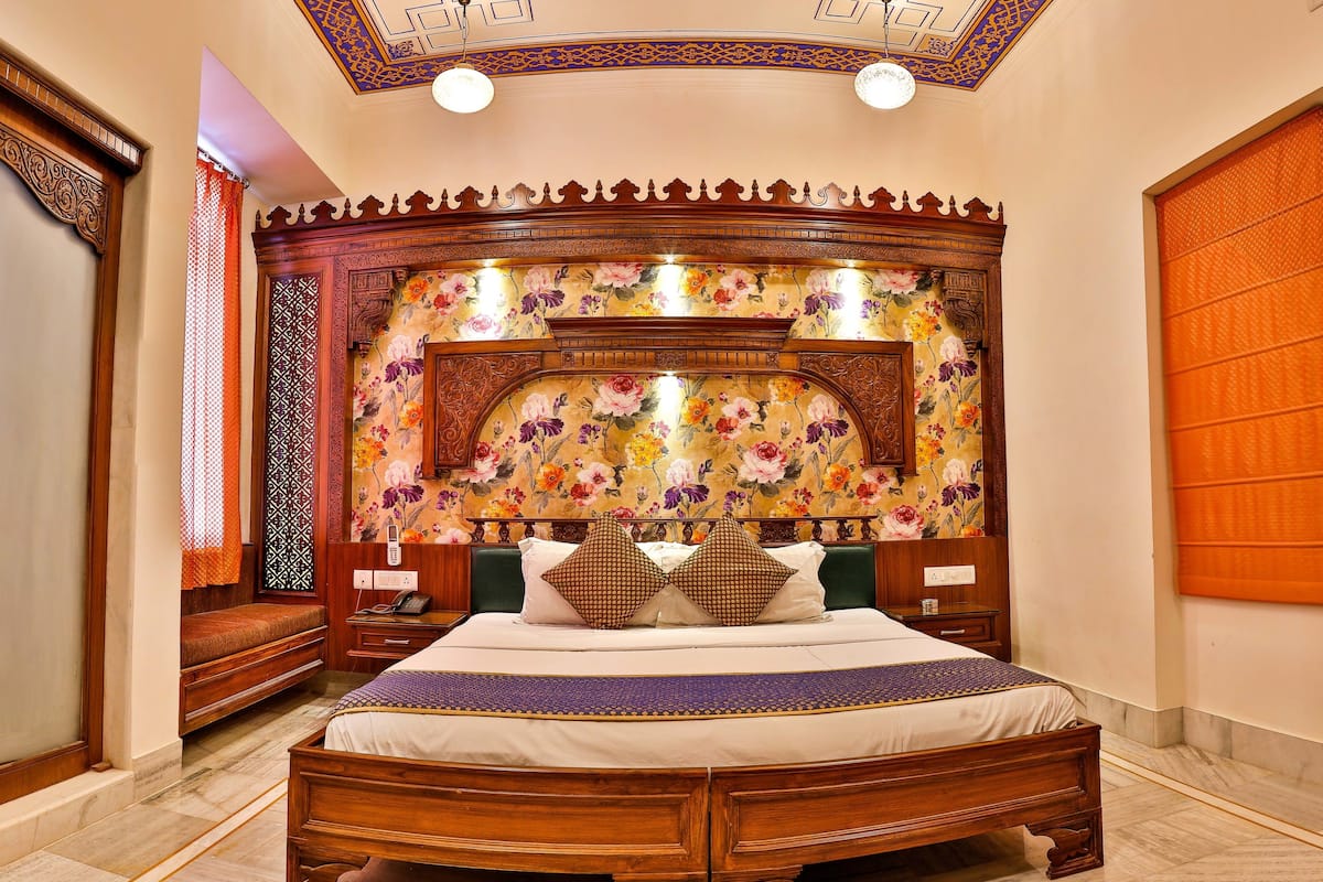 A spacious bedroom with a large bed, ornate wooden headboard, floral wallpaper, and a ceiling with intricate designs.