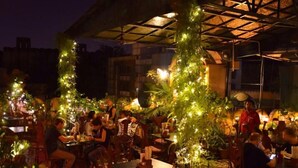 Uma área de refeições ao ar livre com mesas e cadeiras, cercada por vegetação e luzes de Natal.