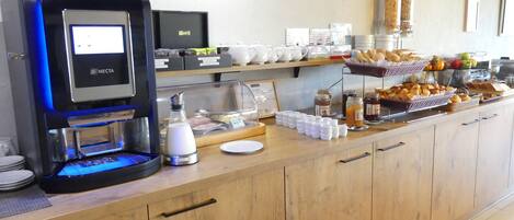 A self-service coffee station with a digital coffee maker, a variety of coffee cups, and a selection of pastries and fruits.