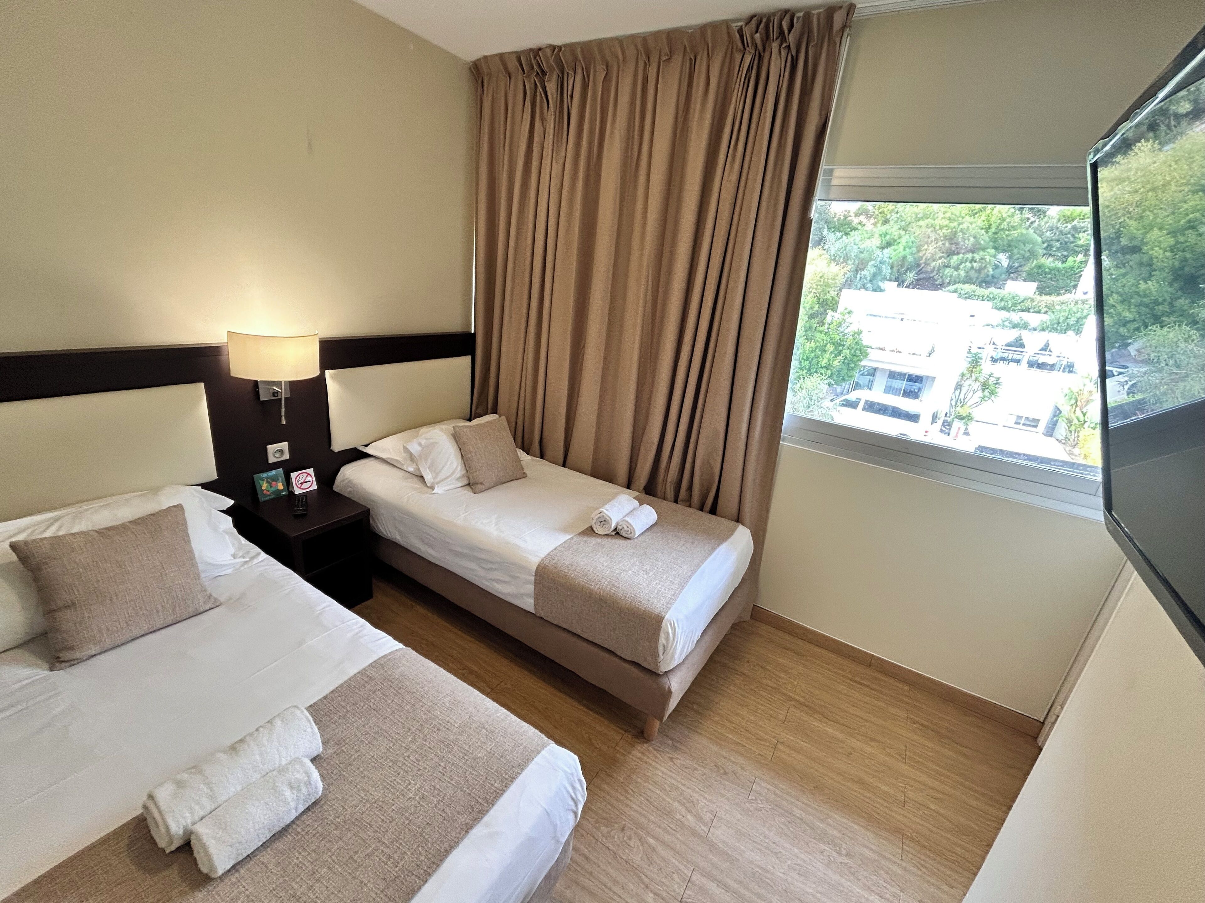 A hotel room with two beds, a nightstand, a lamp, and a window with a view of buildings and trees.