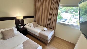 A hotel room with two beds, a nightstand, a lamp, and a window with a view of buildings and trees.