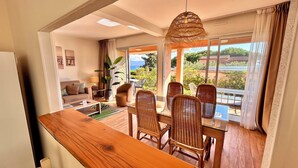 A modern living room with a wooden dining table, wicker chairs, a sofa, and a large window offering a view of the outdoors. - Hôtel La Pinède (Ajaccio)