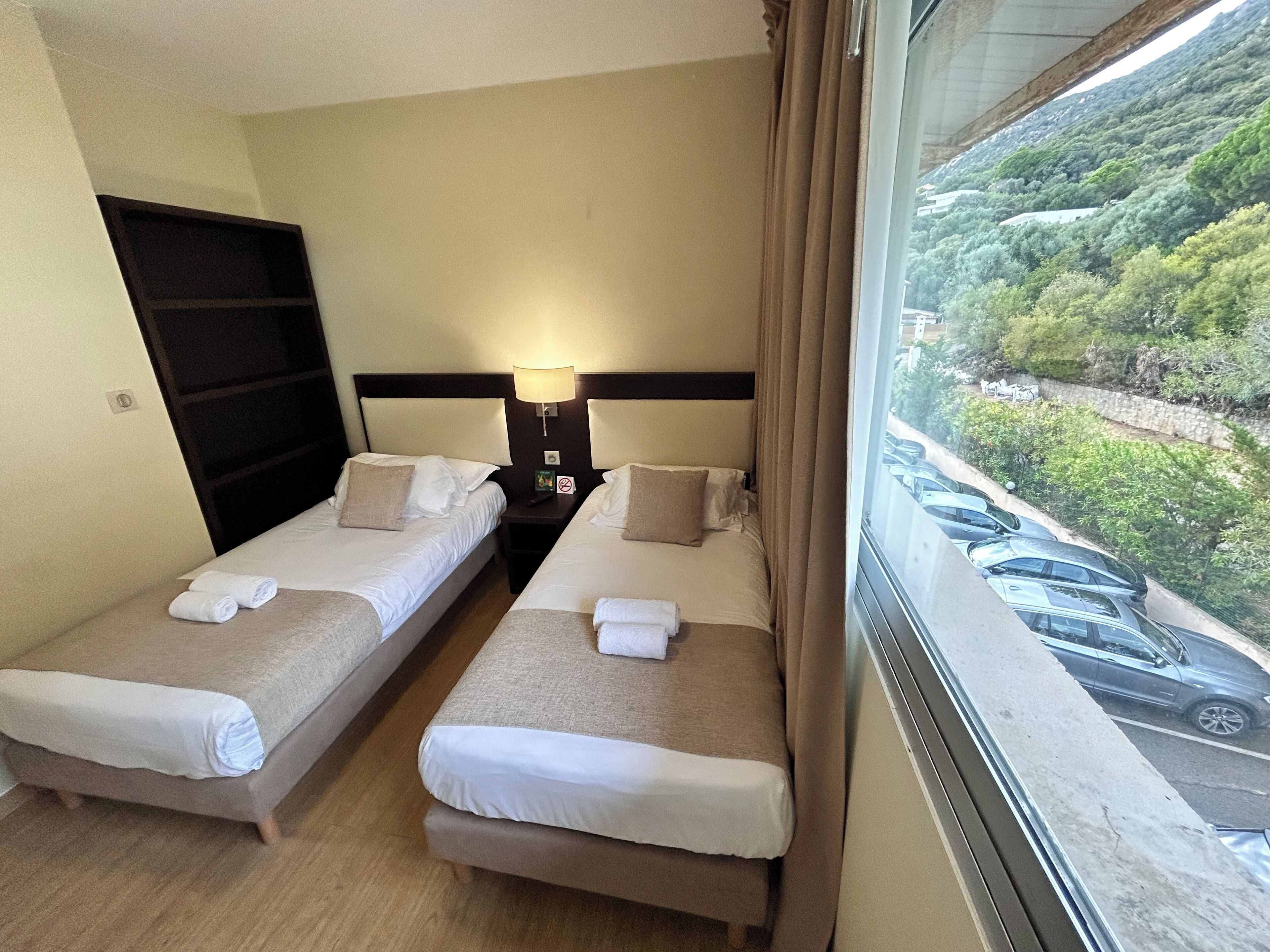 A hotel room with two single beds, a nightstand with a lamp, and a window with a view of parked cars and greenery.