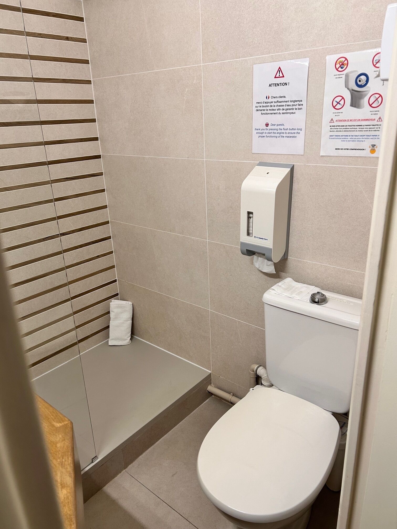 Ein Badezimmer mit Toilette, einem Händetrockner und einem Papierhandtuchspender. An der Wand befinden sich Warnschilder.