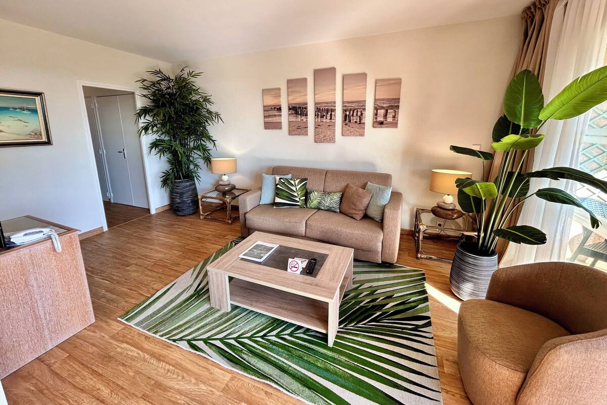 A living room with a sofa, coffee table, and a large plant. There are wall art pieces and a door in the background.