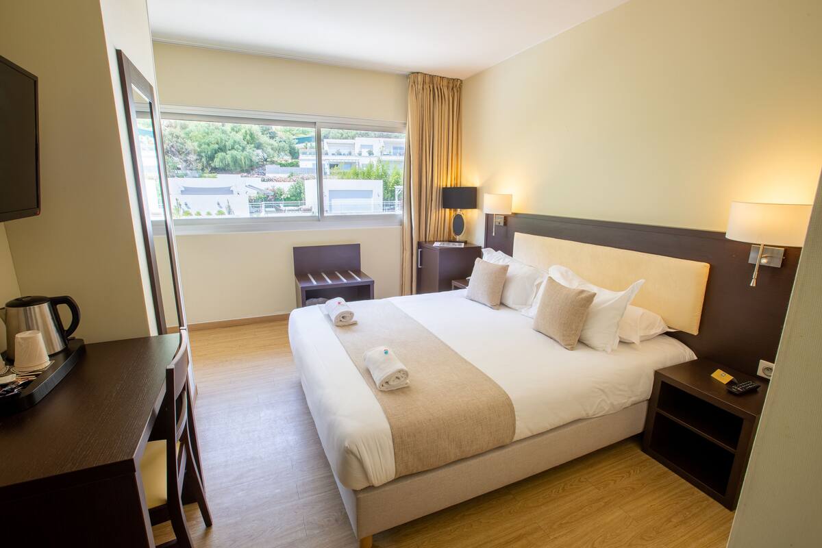 A hotel room with a large bed, bedside tables with lamps, a desk with a coffee maker, and a view of the outside.