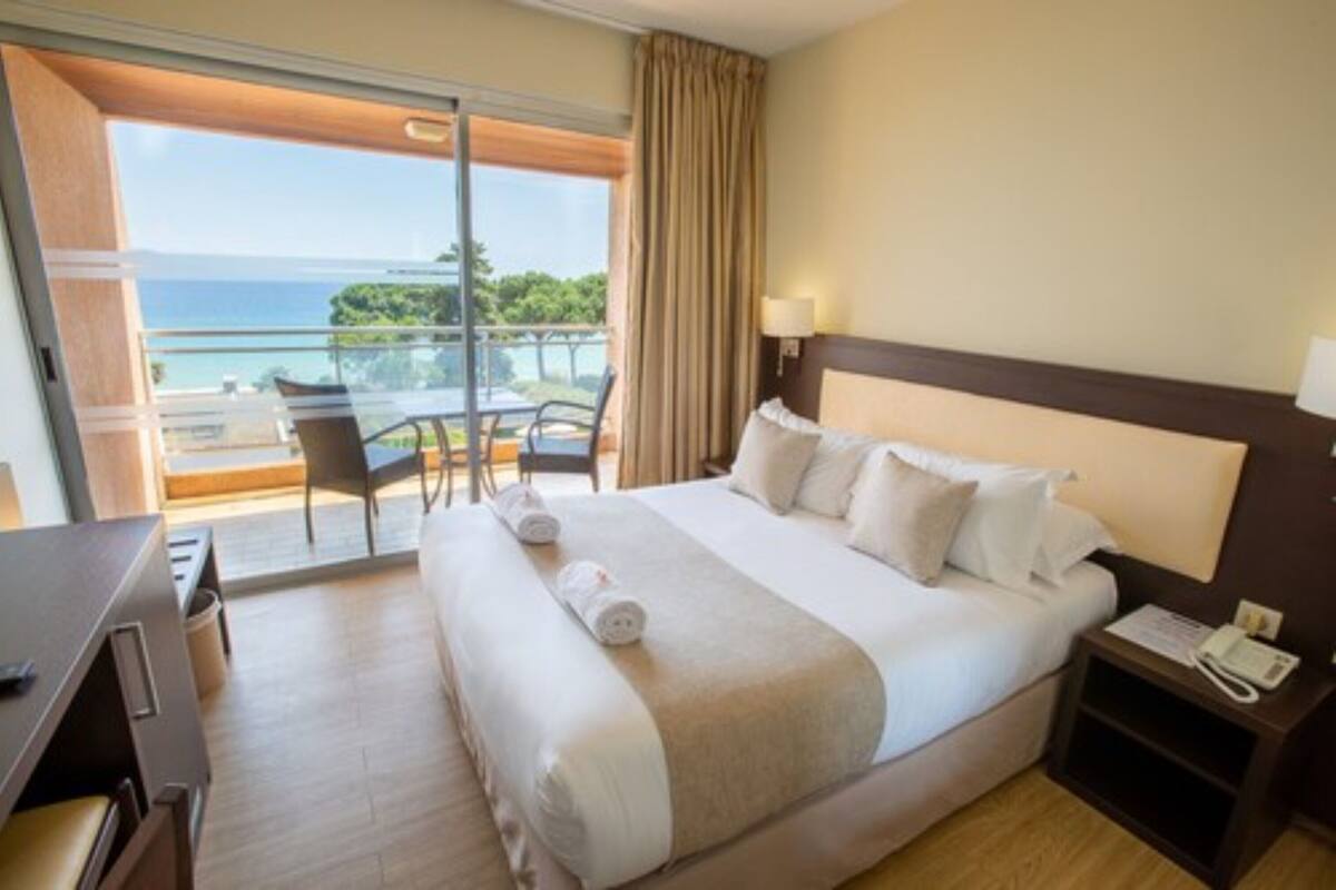 A hotel room with a large bed, a desk, a chair, and a view of the ocean through a sliding glass door.