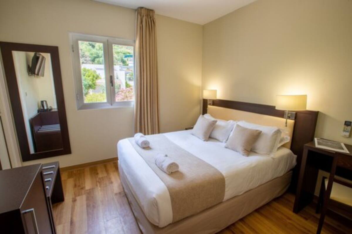 A hotel room with a large bed, a window with curtains, a bedside table with a lamp, and a mirror on the wall.