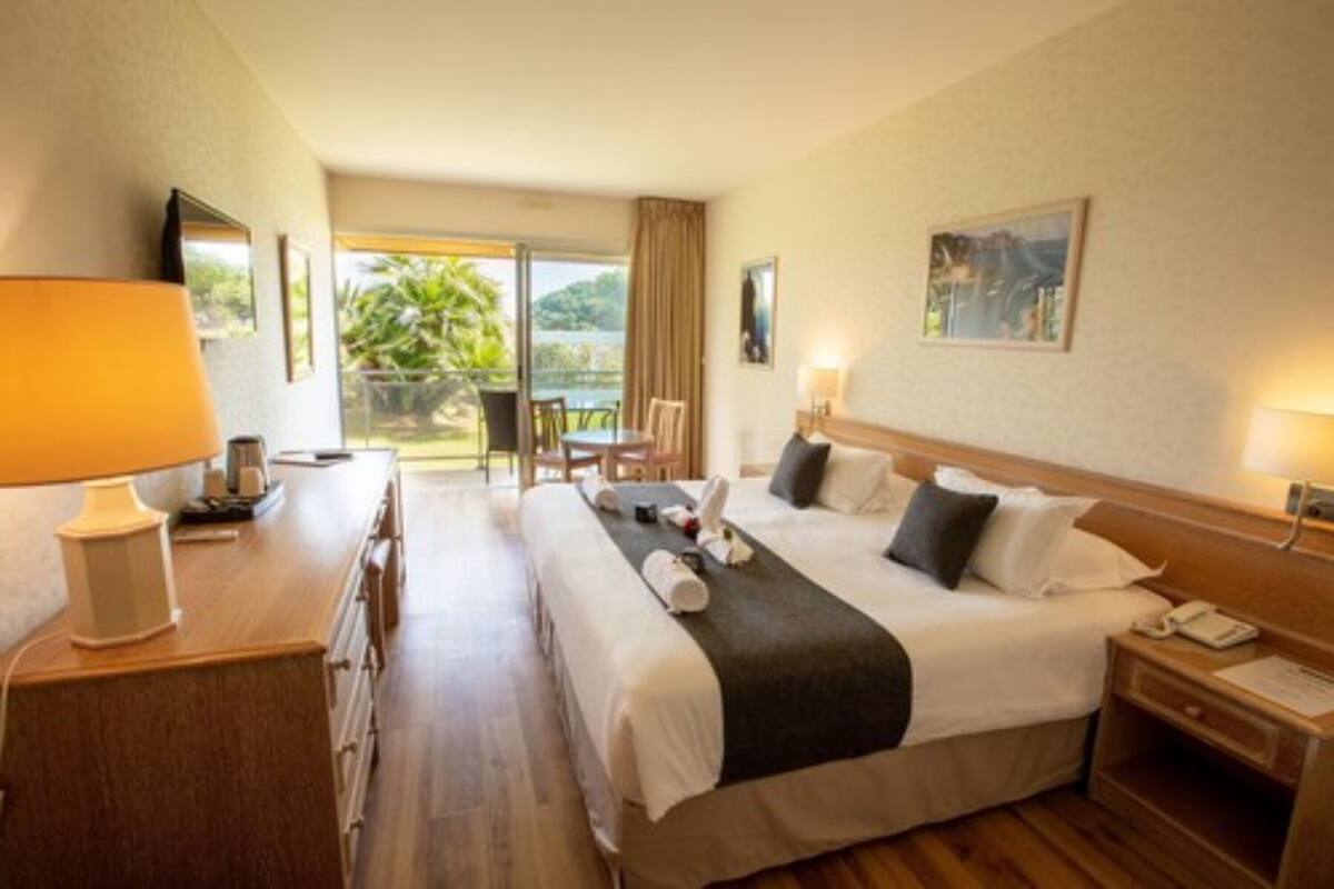 A hotel room with a bed, bedside table, lamp, and a view of the outdoors.