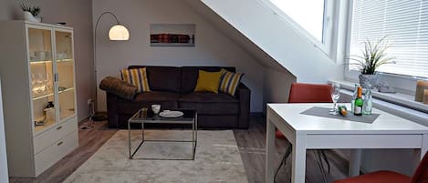 Ein modernes Loft-Apartment mit schrägverdeckter Decke, einem dunkelbraunen Sofa mit gestreiften Kissen, einer Glastruhe, einem weißen Esstisch mit einem roten Stuhl und einem großen Fenster mit Jalousien.