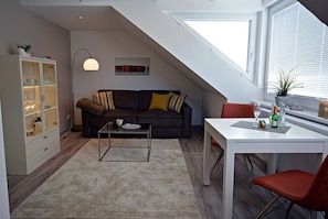 Ein modernes Loft-Apartment mit schrägverdeckter Decke, einem dunkelbraunen Sofa mit gestreiften Kissen, einer Glastruhe, einem weißen Esstisch mit einem roten Stuhl und einem großen Fenster mit Jalousien.