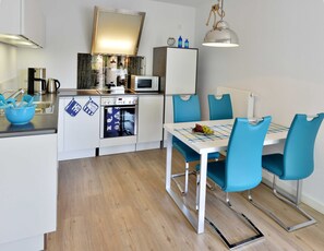 Cozinha compacta com micro-ondas, fogão e pia. Há uma mesa de jantar com cadeiras azuis e toalha de mesa xadrez.