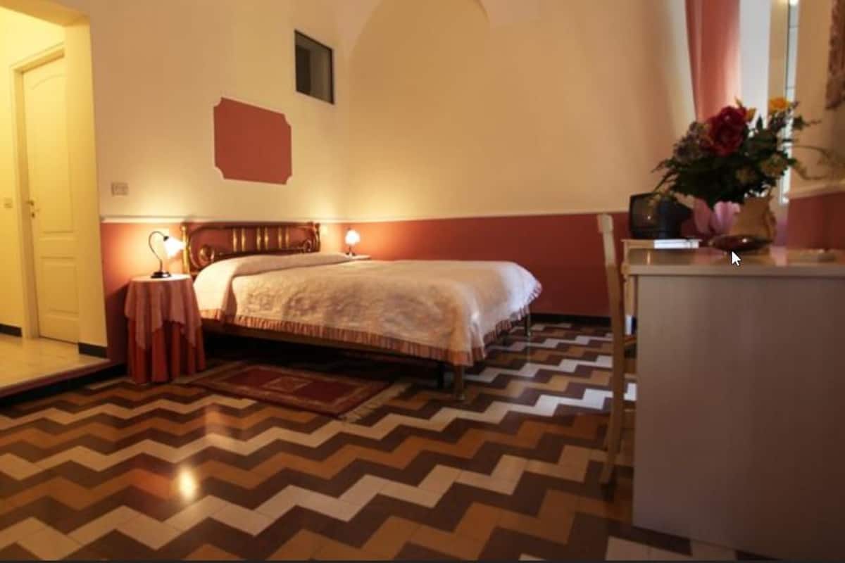 A hotel room with a bed, a nightstand, a desk, and a chair. There is a door and a window with curtains.