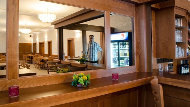 A wooden bar area with a mirror reflecting a man standing in a dining area. - Hotel Pansky dum (Blovice)