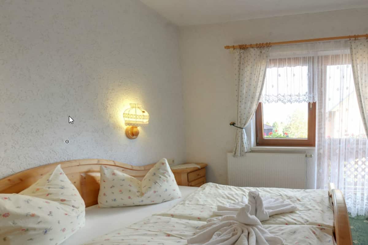A bedroom with a wooden bed, floral bedding, a wall-mounted light, and a window with curtains.