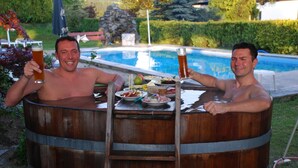 Dois homens dentro de um barril de madeira, segurando copos de cerveja, com uma piscina e vegetação ao fundo.