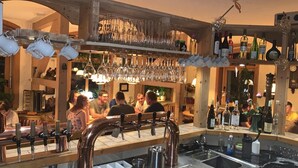 Uma área de bar com várias garrafas de vinho, uma pia e um grupo de pessoas sentadas em mesas.