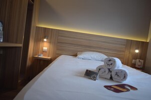Quarto de hotel com cama bem arrumada, mesa de cabeceira com abajur e cabeceira de madeira.