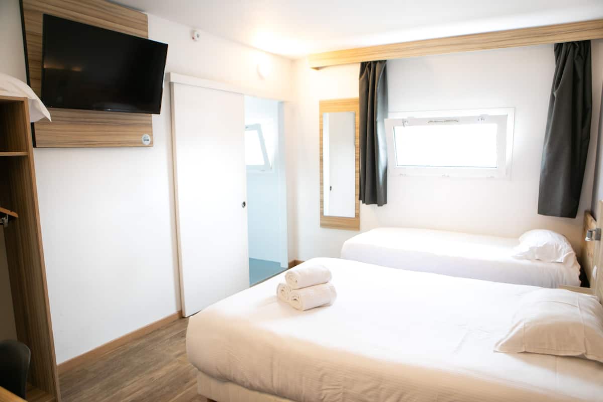 Ein Hotelzimmer mit zwei Betten, einem an der Wand montierten Fernseher, einem Kleiderschrank und einem Fenster mit Vorhängen.