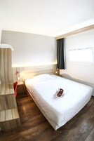 Quarto de hotel com cama, mesa de cabeceira de madeira, uma cadeira vermelha, escrivaninha e janela com cortinas.