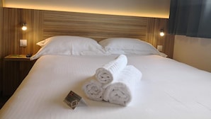 A neatly made bed with white linens and rolled towels, a bedside table with a remote control, and a wall-mounted light fixture. - Best Hotel Bordeaux Sud (Villenave-d'Ornon)