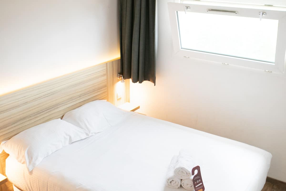 Ein ordentlich bezogenes Bett mit weißen Leinen, ein hölzernes Kopfteil, ein Nachttisch mit Lampe, ein Fenster mit Vorhang und ein Handtuch mit Etikett auf dem Bett.