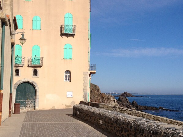 A coastal building with a large green door, multiple windows with green shutters, and a stone wall leading to the sea.