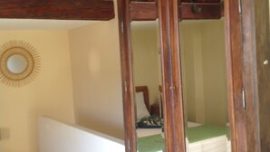 Una habitación con un espejo de marco de madera, una cama con colchón verde y un estante pequeño con libros y una cesta.