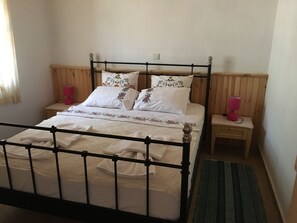 Un lit simple avec un cadre en métal, une literie blanche et des coussins à motifs floraux. Il y a deux tables de chevet avec des lampes, et une tête de lit en bois.