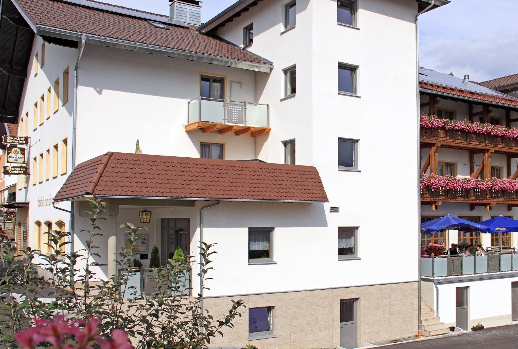 A multi-story building with a white facade, a brown tiled roof, and a balcony with pink flowers.
