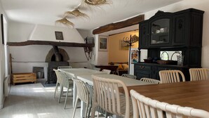 Un coin repas rustique avec une longue table en bois et des chaises blanches, un buffet noir aux portes vitrées et une cheminée en pierre.