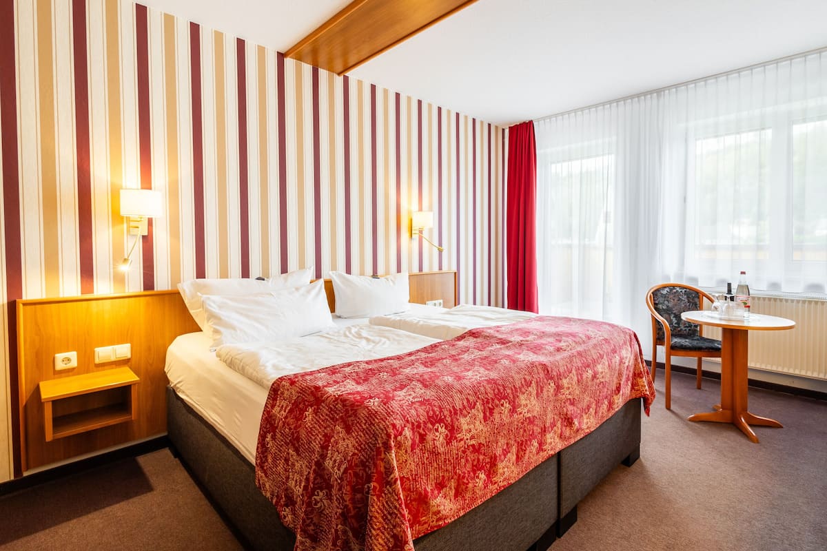 Habitación de hotel con una cama grande, una mesita de noche, una silla, una pequeña mesa redonda y un ventanal con cortinas.