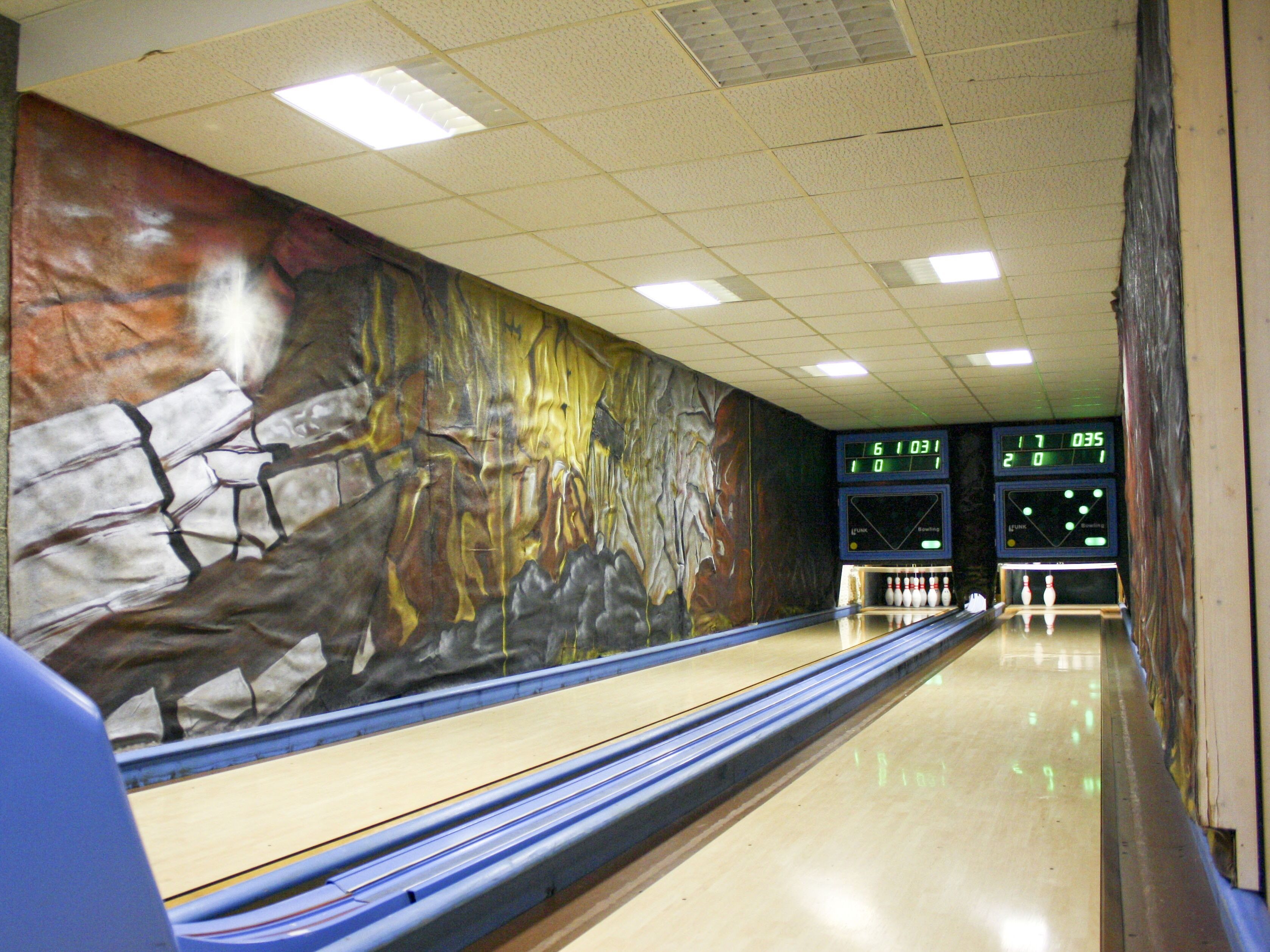 A bowling alley with a mural on the wall and a scoreboard displaying scores.