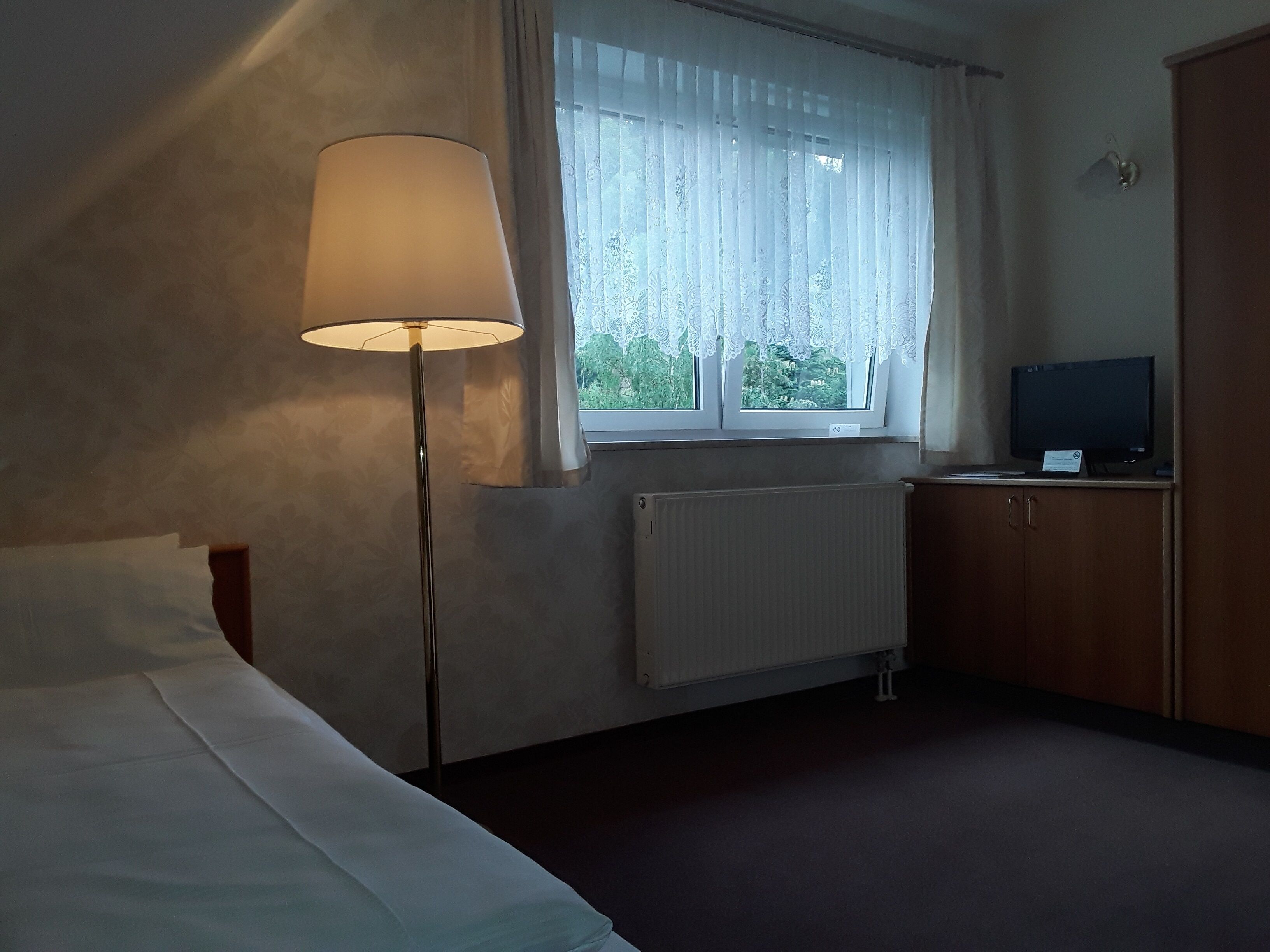 A hotel room with a bed, a standing lamp, a window with sheer curtains, a radiator, and a television on a wooden cabinet.