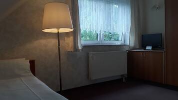 Quarto de hotel com cama, candeeiro, janela com cortinas translúcidas, radiador e televisão sobre um armário de madeira.