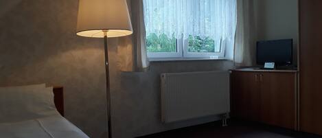 A hotel room with a bed, a standing lamp, a window with sheer curtains, a radiator, and a television on a wooden cabinet.