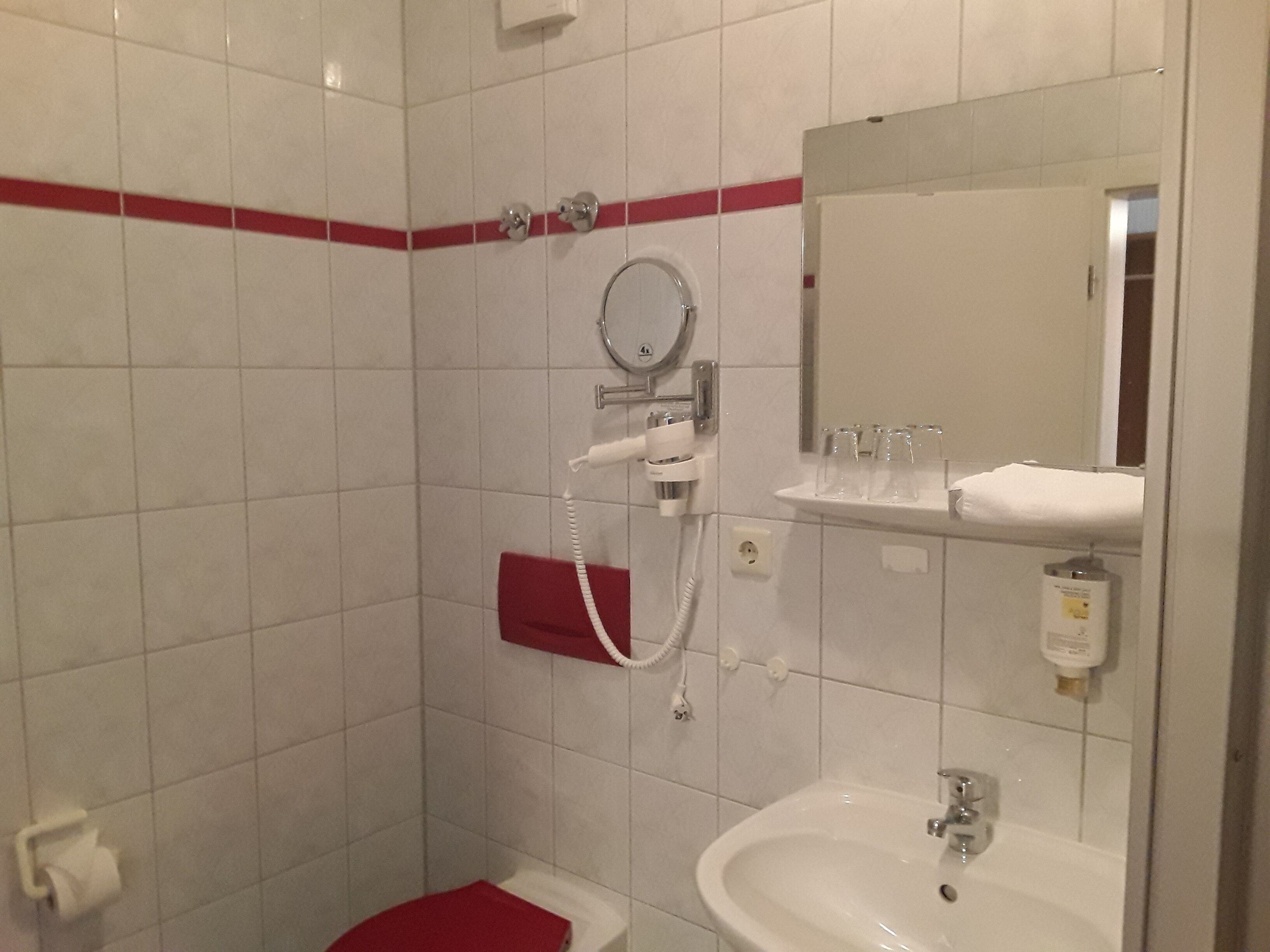 Un baño con un lavabo blanco, un inodoro rojo, una ducha con cabezal de ducha manual y un espejo sobre el lavabo.