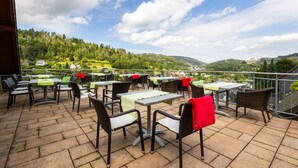 Eine Terrasse im Freien mit Tischen und Stühlen, die auf eine reizvolle Landschaft mit Hügeln und Häusern blickt.