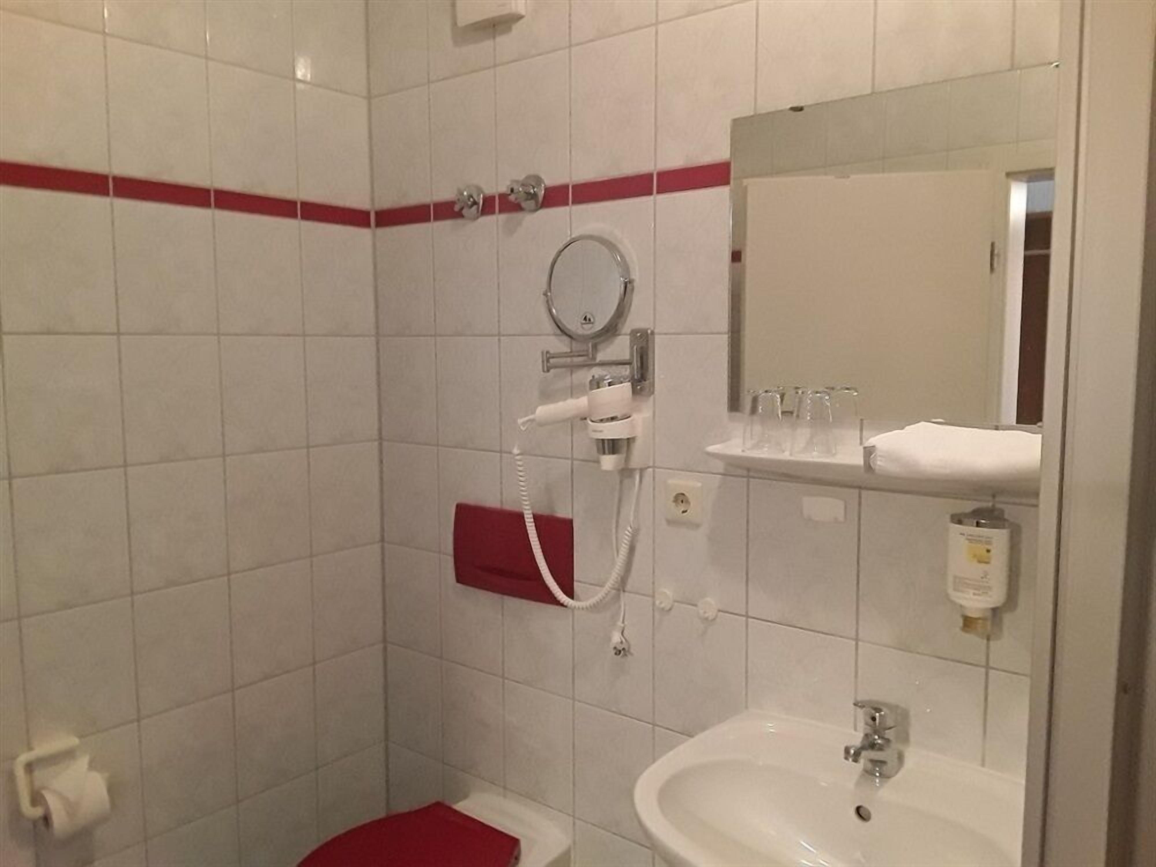 A bathroom with white tiles, a red stripe, a sink, a mirror, a shower, and a toilet.