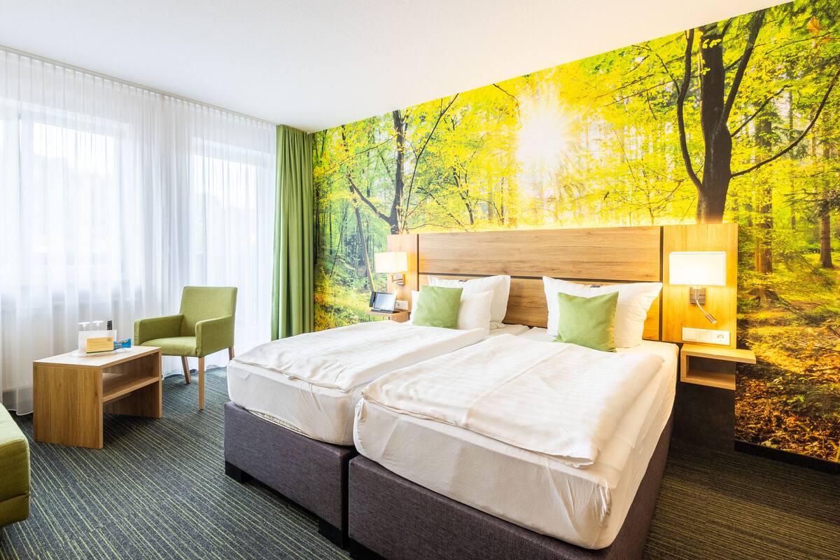 Habitación de hotel con dos camas, un escritorio, una silla y un mural grande con un bosque.