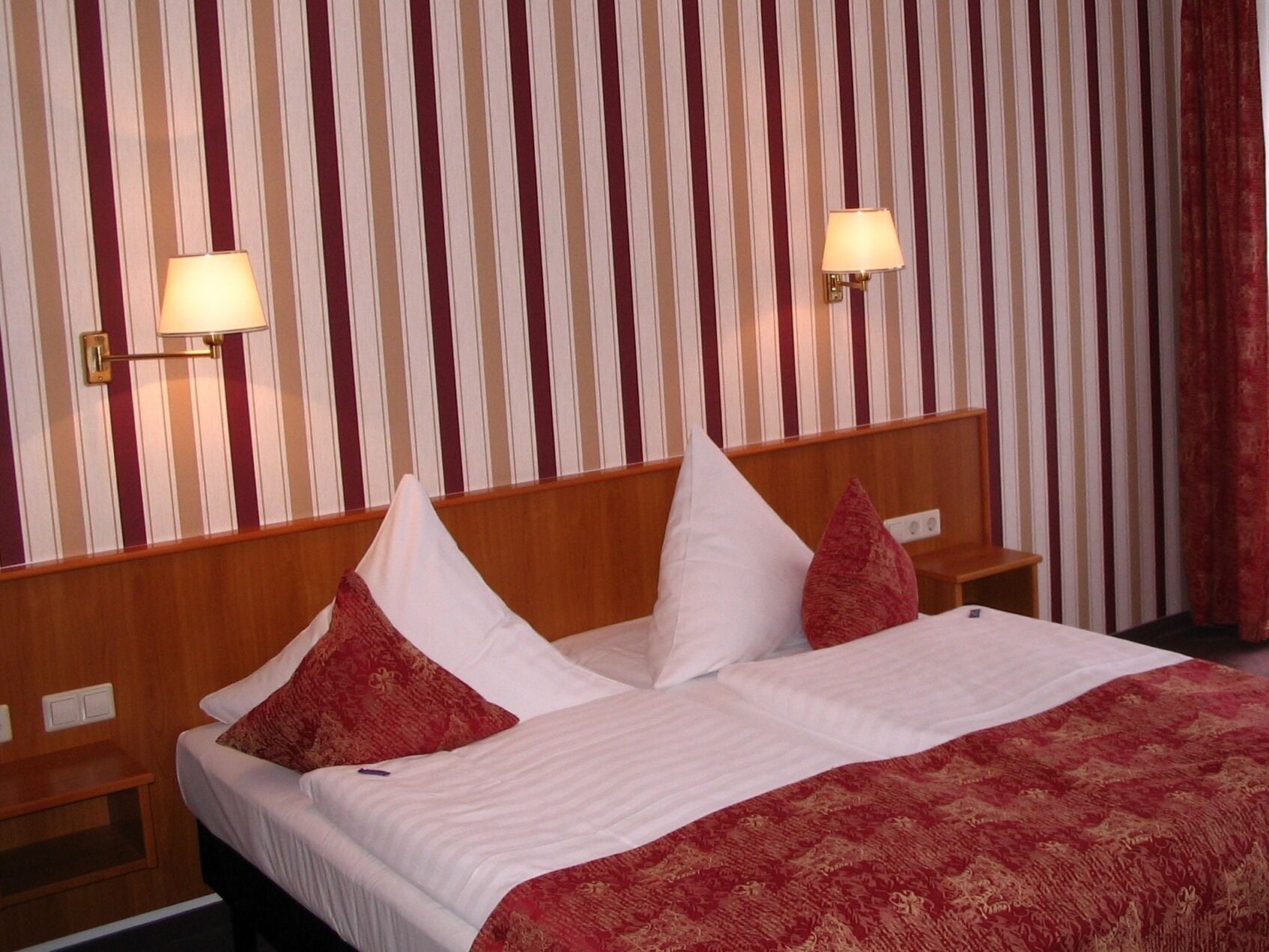 A hotel room with a striped wallpaper, a wooden headboard, two bedside tables with lamps, and a bed with white bedding and red pillows.
